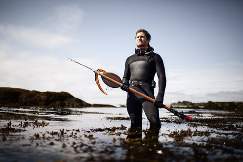 A man in a wetsuit, standing on the shore with a large spear fishing harpoon. - MINF00476