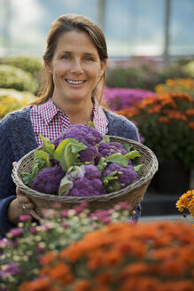 Eine Frau hält eine Schüssel mit frischem Obst und Gemüse, lila sprießender Brokkoli, blühende Pflanzen, Crysanthemen. - MINF00514