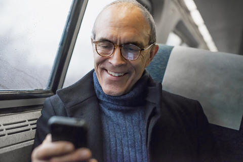 Ein reifer Mann sitzt am Fenster eines Zugwaggons und benutzt sein Mobiltelefon, um unterwegs in Kontakt zu bleiben., lizenzfreies Stockfoto