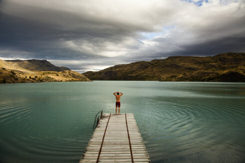 Ein junger Mann steht am Ende eines Holzstegs und bereitet sich darauf vor, in einen ruhigen, von Bergen umgebenen See im Torres del Paine National Park, Chile, zu springen. - MINF00664