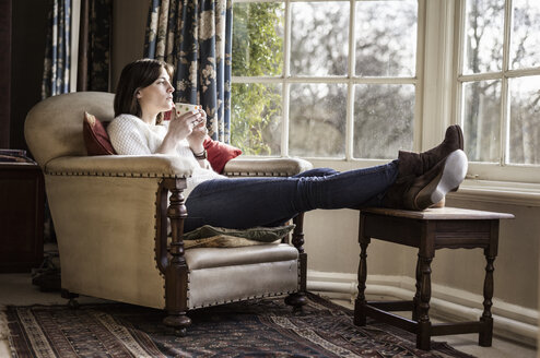 Eine junge Frau entspannt sich zu Hause, legt die Füße hoch und trinkt eine Tasse Tee. - MINF00975