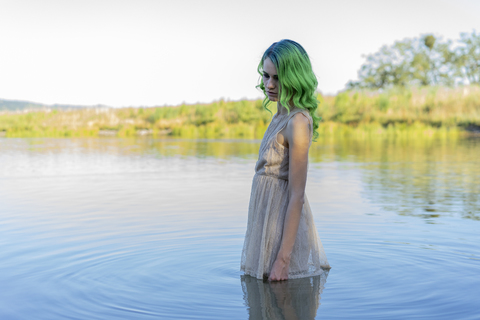 Junge Frau mit gefärbten grünen Haaren steht bekleidet im Wasser eines Sees, lizenzfreies Stockfoto