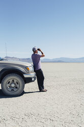 Mann steht in der Wüste, schaut durch ein Fernglas und lehnt sich an einen Truck, Black Rock Desert - MINF01049