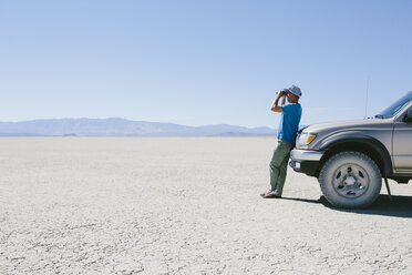 Mann steht in der Wüste, schaut durch ein Fernglas und lehnt sich an einen Truck, Black Rock Desert - MINF01050