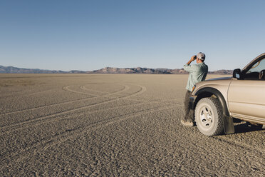 Mann steht in der Wüste, schaut durch ein Fernglas und lehnt sich an einen Truck, Black Rock Desert - MINF01055