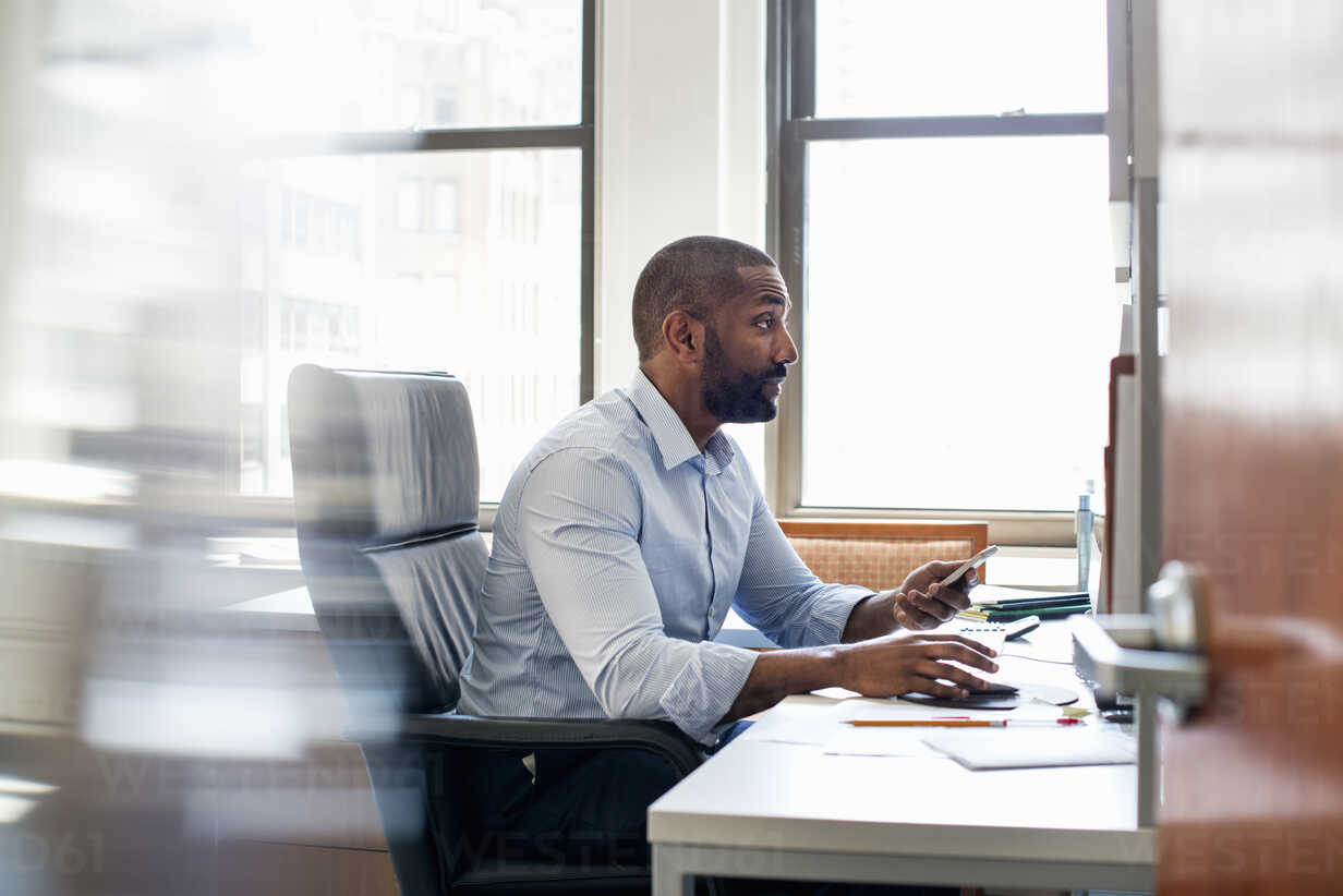 A man working in an office holding his smart phone and checking his  computer. stock photo