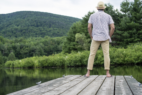 Ein Mann steht auf einem Holzsteg und blickt auf einen ruhigen See., lizenzfreies Stockfoto