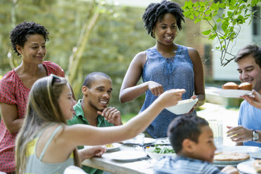 Eine Familie versammelt sich, Männer, Frauen und Kinder um einen Tisch in einem Garten im Sommer. - MINF01274