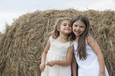 Two girls standing by a large haybale, smiling and sharing a secret. - MINF01299