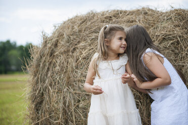 Two girls standing by a large haybale, smiling and sharing a secret. - MINF01300