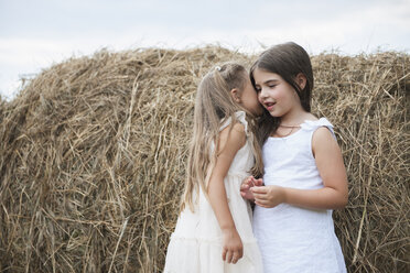 Two girls standing by a large haybale, smiling and sharing a secret. - MINF01301