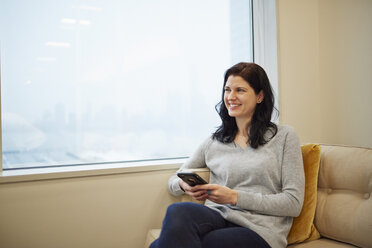 Eine Geschäftsfrau sitzt an einem Fenster und hält ein Smartphone in der Hand. - MINF01312
