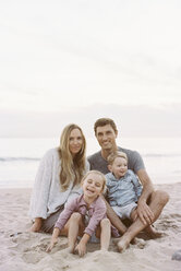 Ein Ehepaar sitzt mit seinem Sohn und seiner Tochter an einem Sandstrand am Meer, schaut in die Kamera und lächelt. - MINF01393