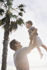 Man standing by a palm tree, playing with his young son, lifting him into the air. - MINF01398