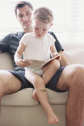 Mann sitzt mit seinem Sohn auf einem Sofa, hält ein Buch in der Hand und liest. - MINF01402