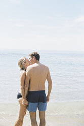 Couple in swimwear standing on a sandy beach by the ocean, kissing. - MINF01411