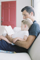Man sitting on a sofa with his son sitting on his lap, holding a book, reading a story. - MINF01431