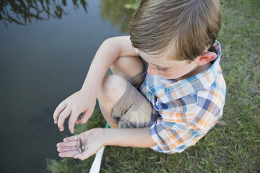 A young boy outdoors sitting on a riverbank with a small fish in the palm of his hand. - MINF01525