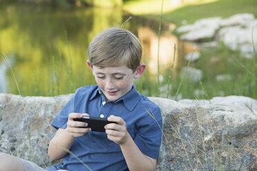 Ein kleiner Junge sitzt im Freien an einen Felsen gelehnt und spielt ein elektronisches Handheld-Spiel. - MINF01530