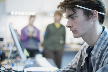 A young man seated at a computer in a repair shop. Two people in the background. - MINF01579