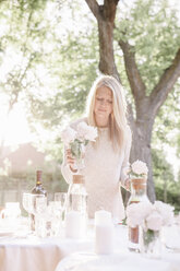 Blonde Frau deckt einen Tisch in einem Garten, Kerzen und Vasen mit rosa Rosen. - MINF01649