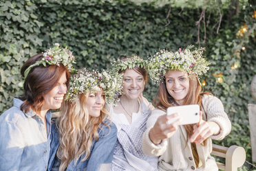 Vier lächelnde Frauen mit Blumenkränzen im Haar, die in einem Garten sitzen und ein Selfie machen. - MINF01650