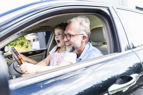 Kleines Mädchen sitzt auf dem Schoß des Großvaters und tut so, als ob sie das Auto lenken würde - UUF14571