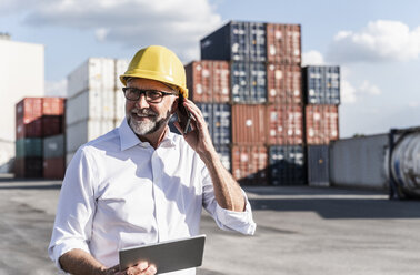 Geschäftsmann im Frachthafen, trägt einen Schutzhelm und benutzt ein Smartphone und ein digitales Tablet - UUF14591