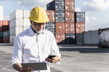 Geschäftsmann im Frachthafen, trägt einen Schutzhelm und benutzt ein Smartphone und ein digitales Tablet - UUF14592
