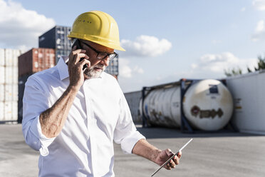 Geschäftsmann im Frachthafen, trägt einen Schutzhelm und benutzt ein Smartphone und ein digitales Tablet - UUF14593