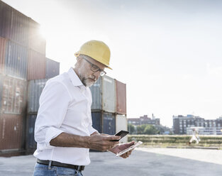 Businessman at cargo harbour, wearing safety helmet, using smartphone and digital tablet - UUF14594