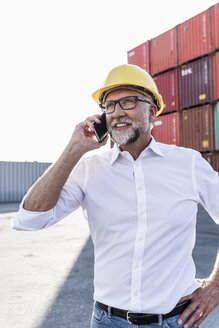 Geschäftsmann im Frachthafen, trägt einen Schutzhelm und benutzt ein Smartphone - UUF14606