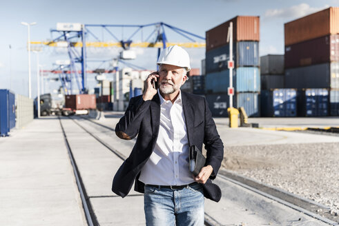 Businessman at cargo harbour, wearing safety helmet, using smartphone - UUF14611