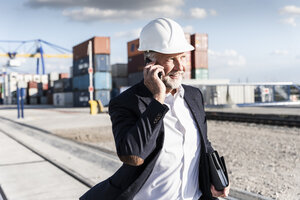 Geschäftsmann im Frachthafen, trägt einen Schutzhelm und benutzt ein Smartphone - UUF14613