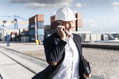 Businessman at cargo harbour, wearing safety helmet, using smartphone - UUF14613