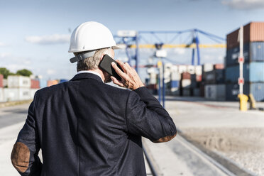 Businessman at cargo harbour, wearing safety helmet, using smartphone - UUF14614