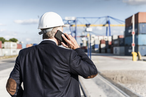 Geschäftsmann im Frachthafen, trägt einen Schutzhelm und benutzt ein Smartphone - UUF14614