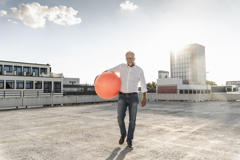 Mature man playing with orange fitness ball on rooftop of a high-rise building stock photo