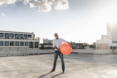 Älterer Mann spielt mit orangefarbenem Fitnessball auf dem Dach eines Hochhauses - UUF14632