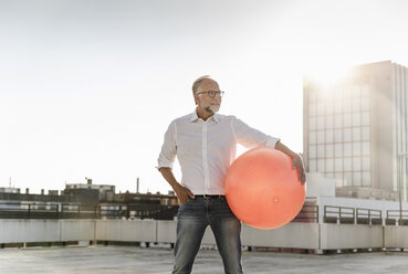 Älterer Mann spielt mit orangefarbenem Fitnessball auf dem Dach eines Hochhauses - UUF14633