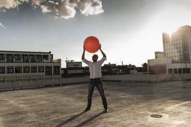 Mature man playing with orange fitness ball on rooftop of a high-rise building - UUF14634