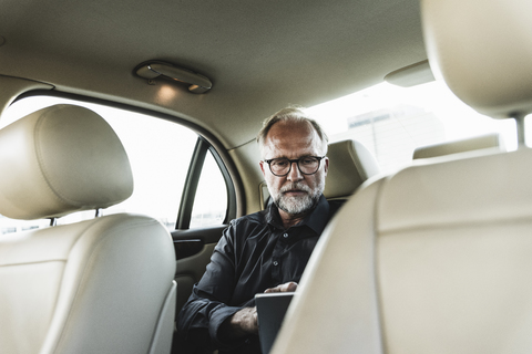 Älterer Geschäftsmann sitzt auf der Rückbank im Auto und benutzt ein digitales Tablet, lizenzfreies Stockfoto