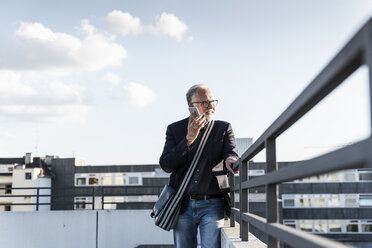 Mature man standing on rooftop, leaning on railing, using smartphone - UUF14652