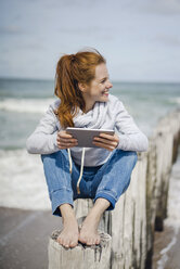 Woman sitting on fence at the beach,using digital tablet at the sea - KNSF04213