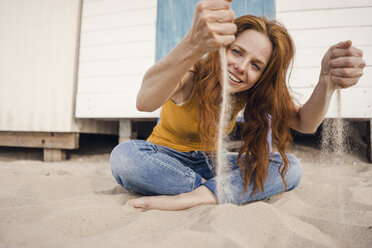Rothaarige Frau, die vor einer Strandhütte sitzt und Sand durch ihre Hände rieseln lässt - KNSF04256