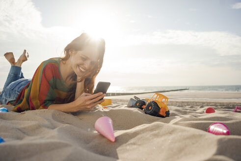 Redheaded woman lying on the beach with beach toys, using smartphone - KNSF04262