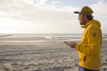 Mann in gelber Jacke, mit Smartphone am Strand - KNSF04271