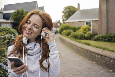 Rothaarige Frau mit Kopfhörern und Smartphone in der Stadt, lizenzfreies Stockfoto