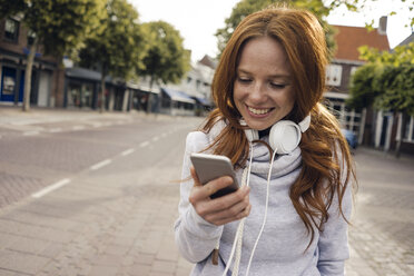 Redheaded woman using headphones and smartphone in the city - KNSF04287