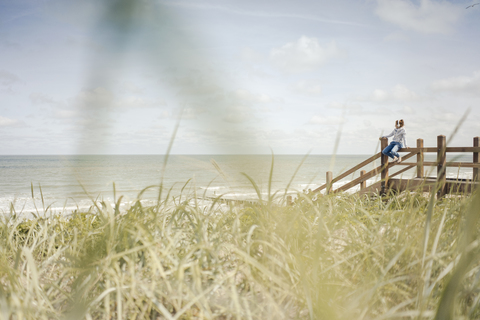 Frau sitzt auf einem Zaun am Meer und trägt Kopfhörer, lizenzfreies Stockfoto